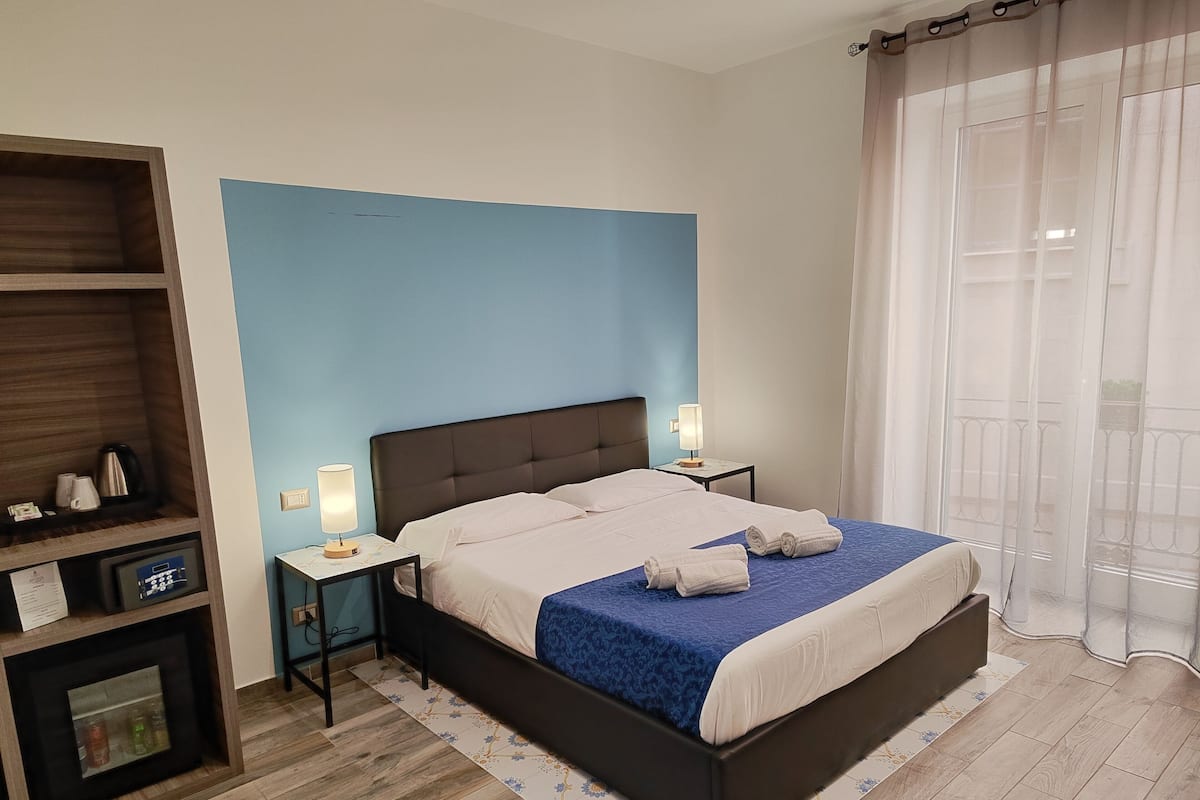 Una habitación de hotel moderna con una cama grande, dos mesitas de noche con lámparas, un estante de madera y vistas al exterior a través de una ventana con cortinas translúcidas.
