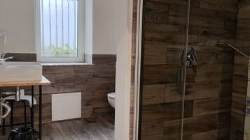 Een badkamer met een houten vloer, een douchecabine van glas, een raam met zonwering en een toilet.