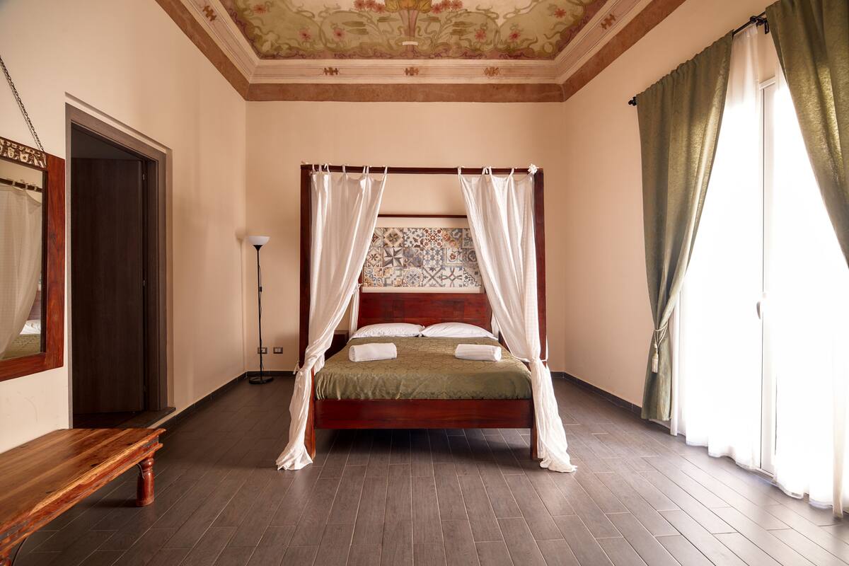 Una cama con dosel y cuatro columnas, un banco de madera, una lámpara de pie y un papel tapiz estampado.