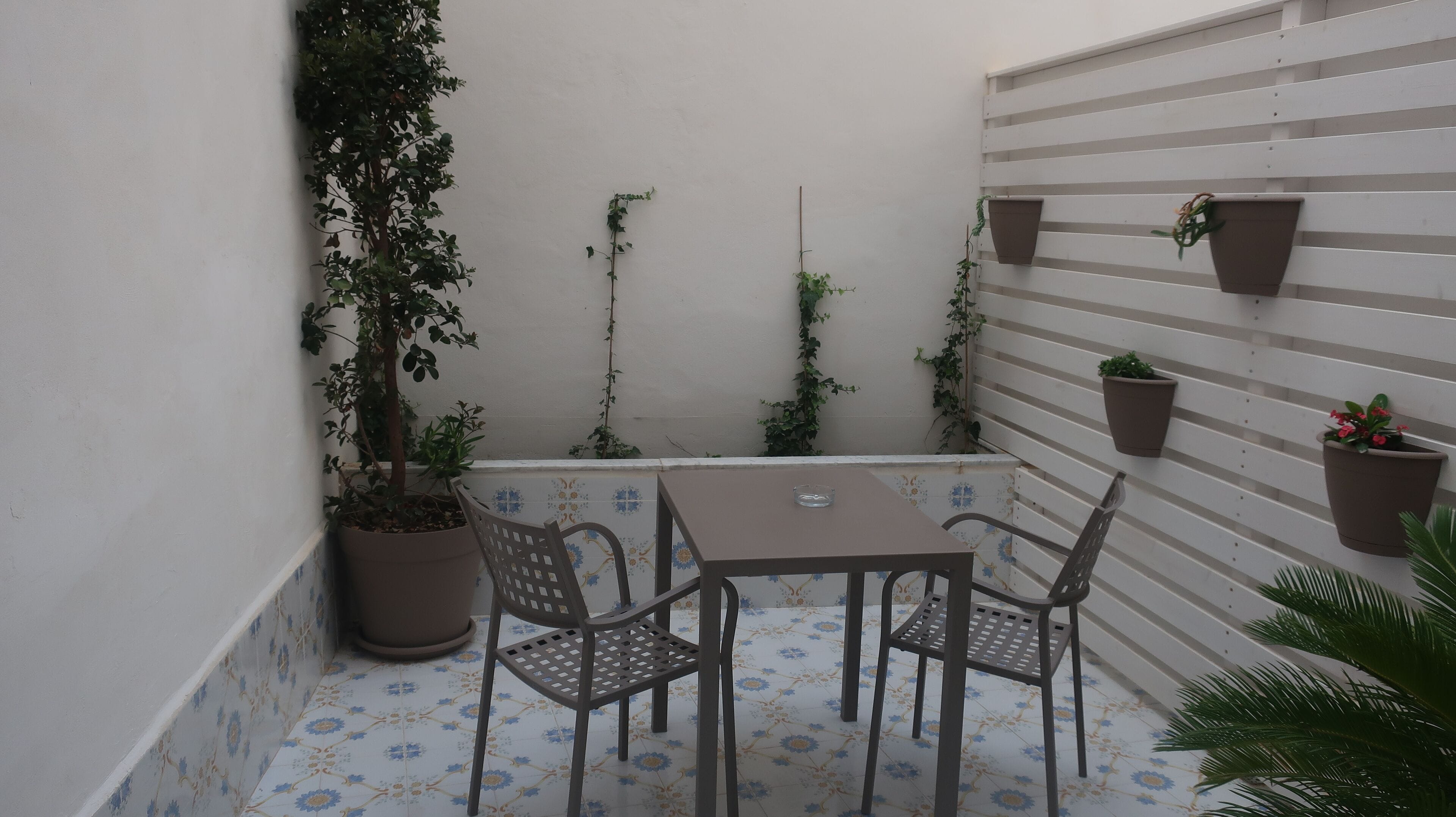 Eine kleine Terrasse im Freien mit Tisch und Stühlen, umgeben von Blumentöpfen und einem gefliesten Boden.