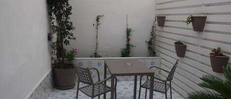 A small outdoor patio with a table and chairs, surrounded by potted plants and a tiled floor.