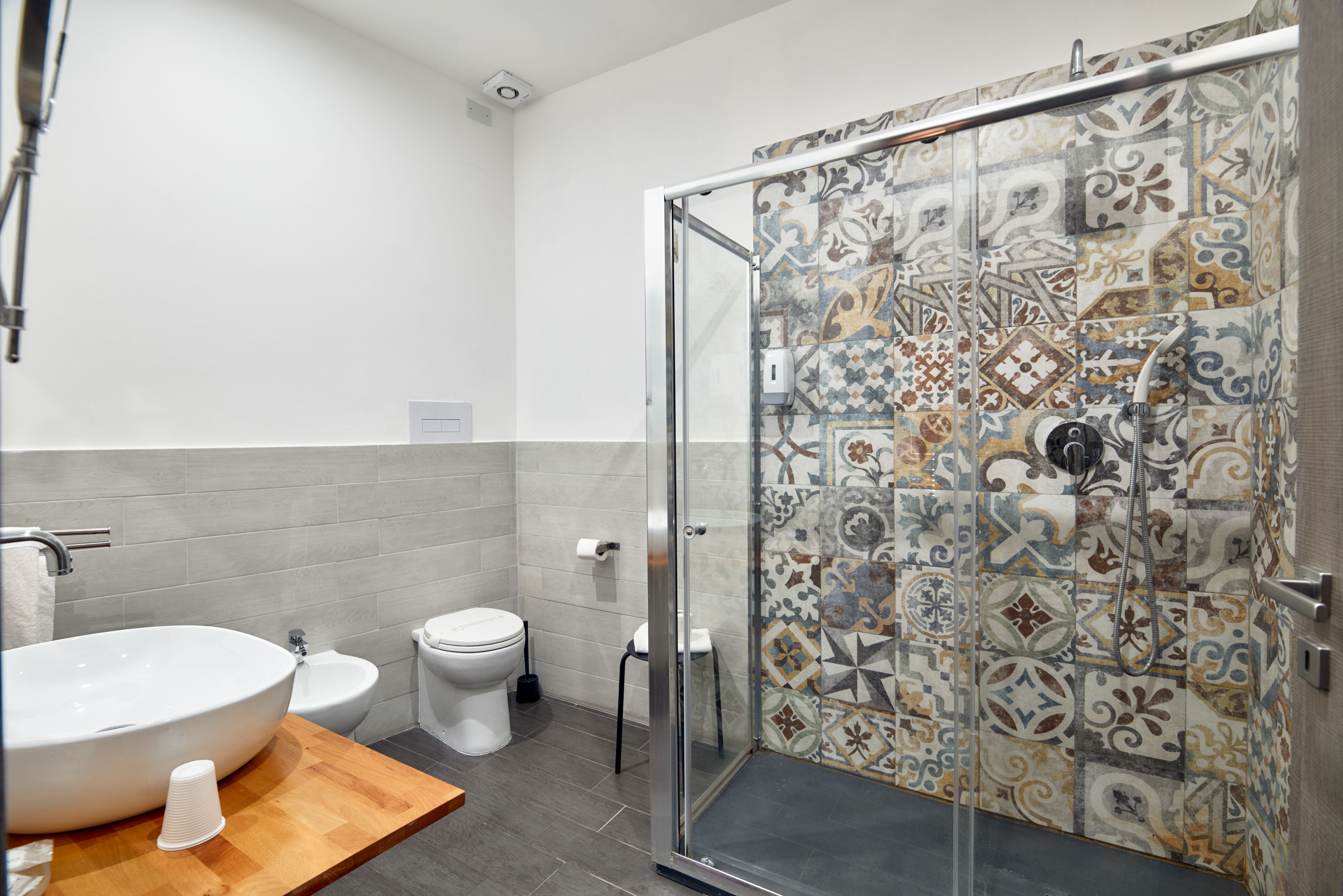 A modern bathroom with a large white sink, a bidet, a toilet, and a shower with patterned tiles.