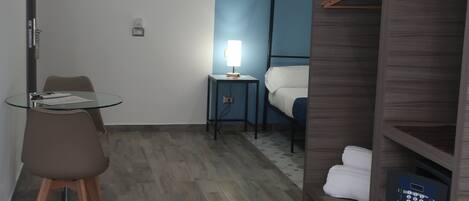 Una habitación de hotel con cama, escritorio con silla, armario y una pequeña mesa con lámpara.