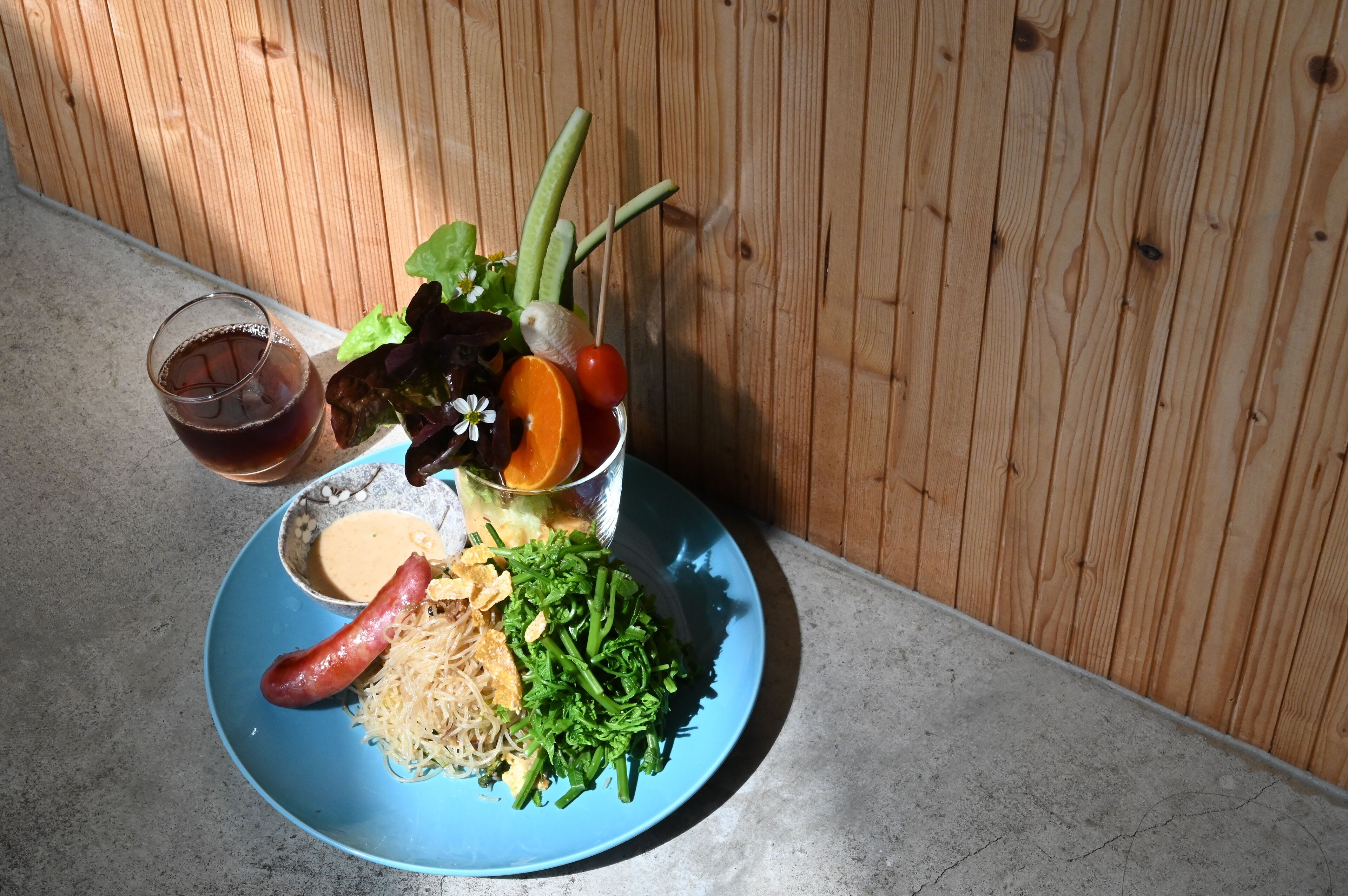 Un plato con fideos, verduras verdes, una salchicha y un vaso de líquido oscuro.