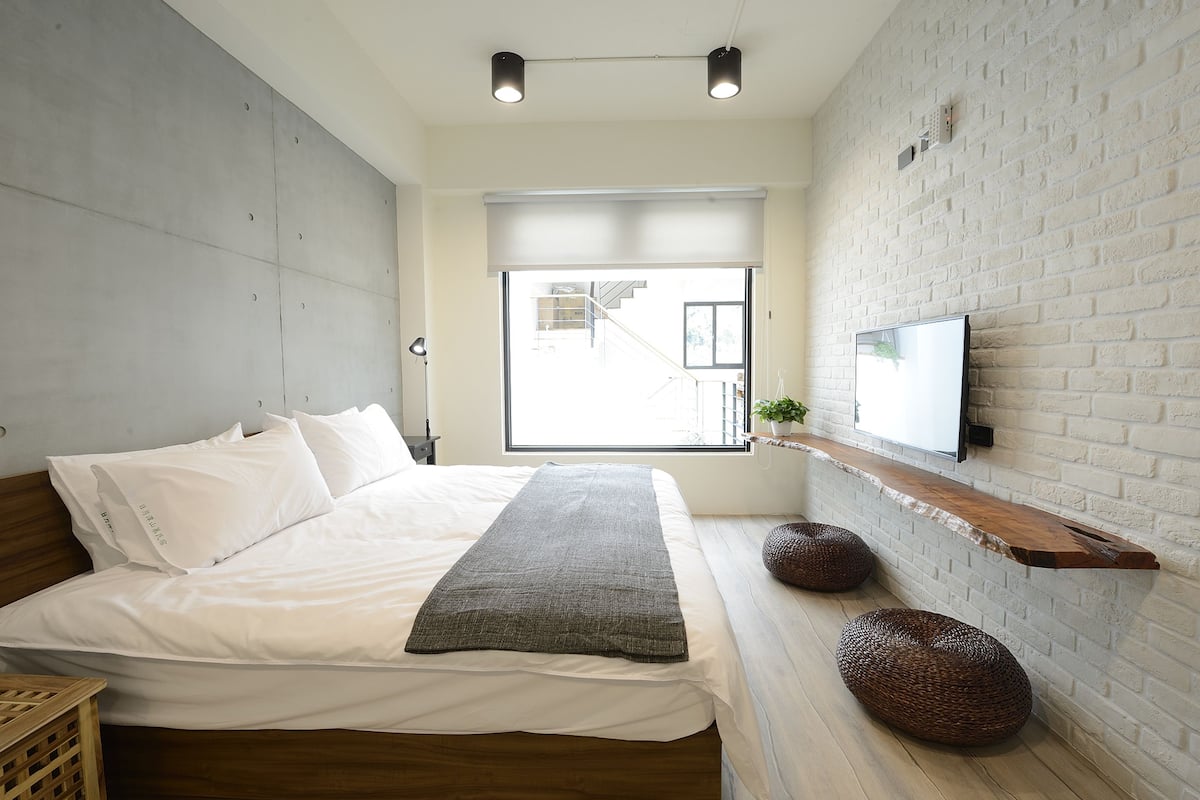 A modern bedroom with a large bed, a TV mounted on a brick wall, and a window with a view of the outside.