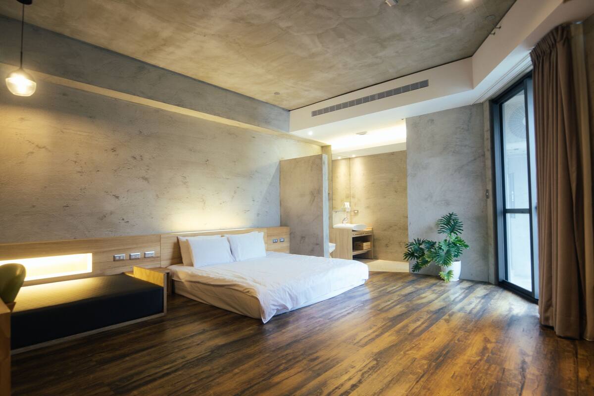 A modern hotel room with a large bed, wooden flooring, a built-in bench, a bedside table, a potted plant, a large window with curtains, and a bathroom with a sink and mirror.