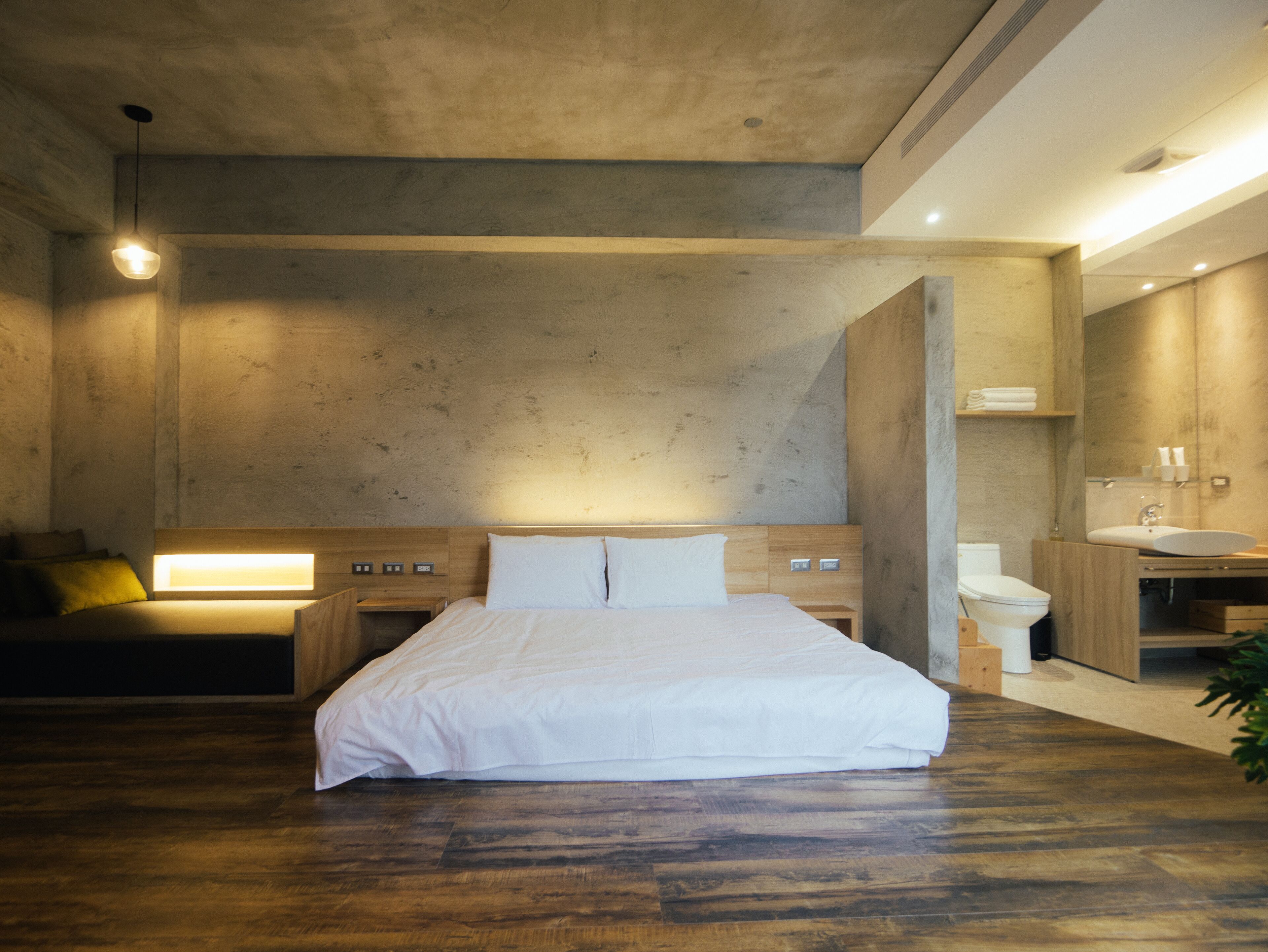 Habitación de hotel moderna con una cama grande, cabecera de madera, mesita de noche y un banco empotrado. Incluye un espejo grande, lavabo e inodoro.