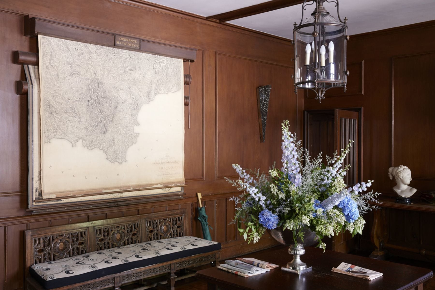 A room with a large map on the wall, a wooden bench, a vase with flowers, and a bust on a table.