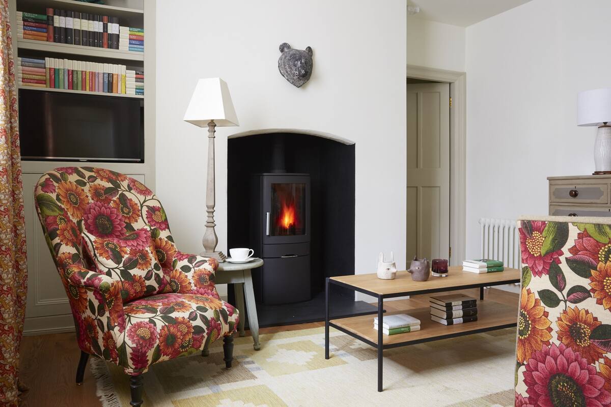 Una sala de estar acogedora con chimenea, un sillón estampado, una mesita de noche con lámpara y una estantería llena de libros.
