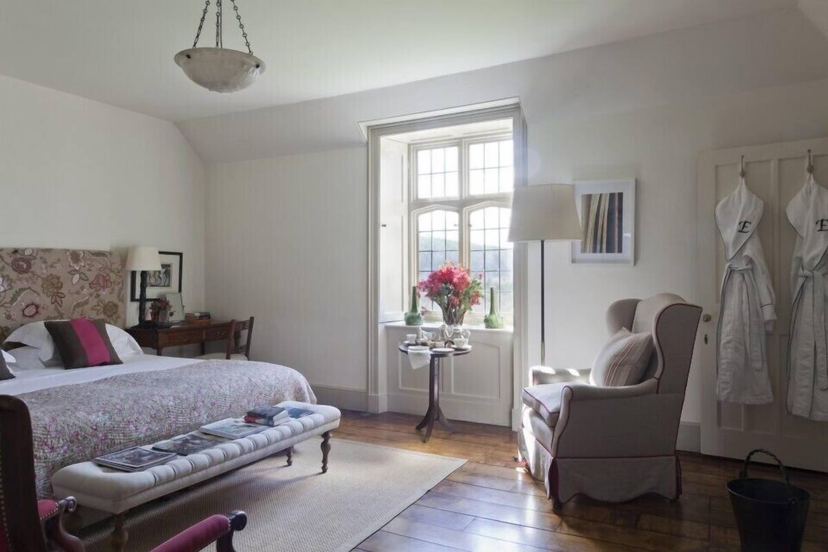 Un dormitorio con una cama, una silla, una mesa con flores y una ventana.