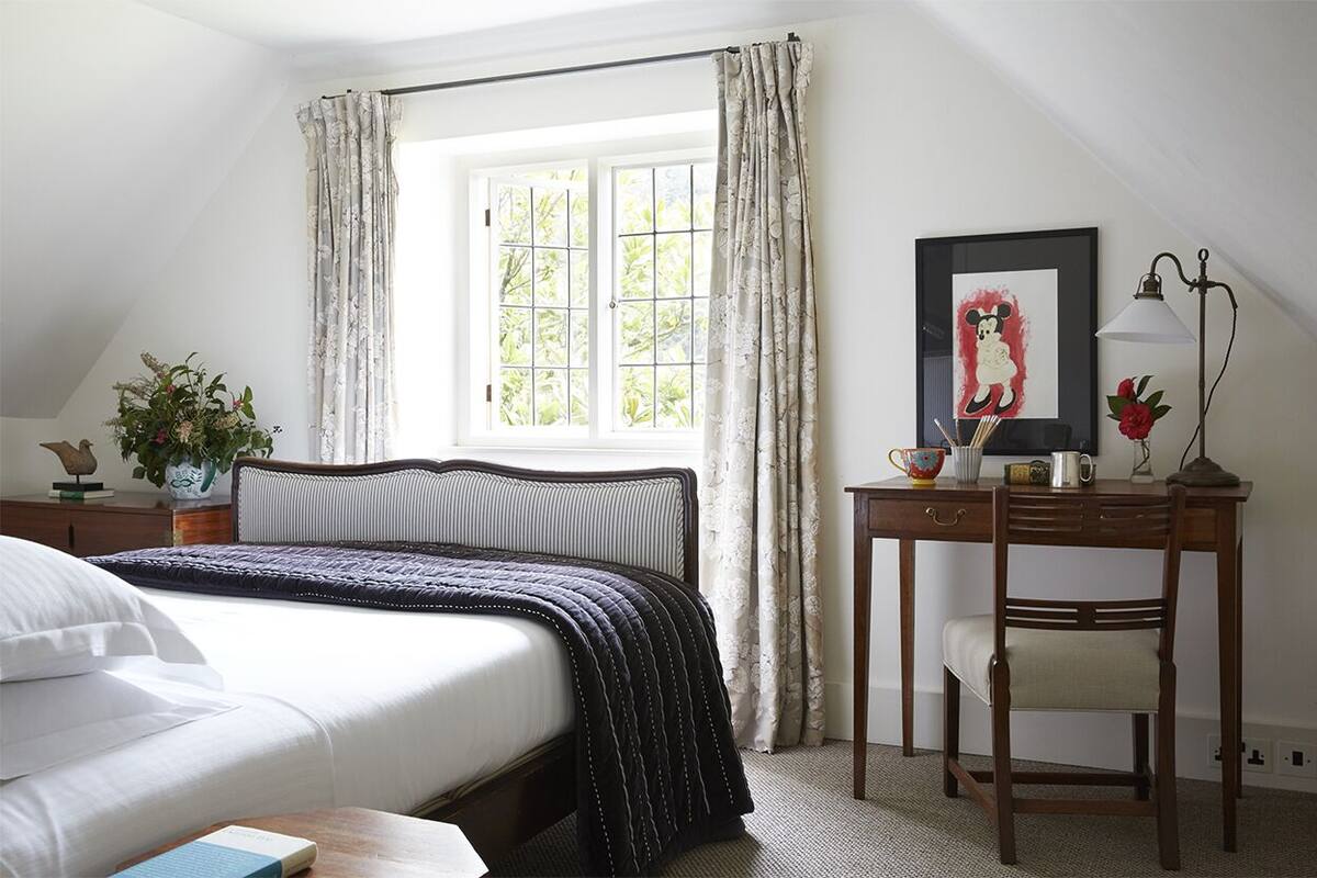 Un dormitorio con cama, escritorio, silla, ventana con cortinas, lámpara y un cuadro en la pared.