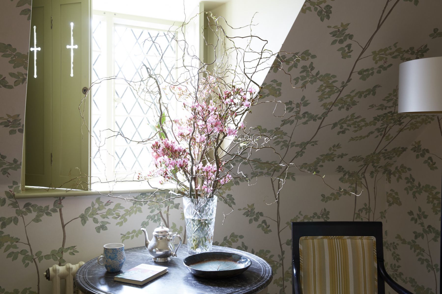 A room with a table set for tea, a vase with pink flowers, a teapot, a cup, and a plate.