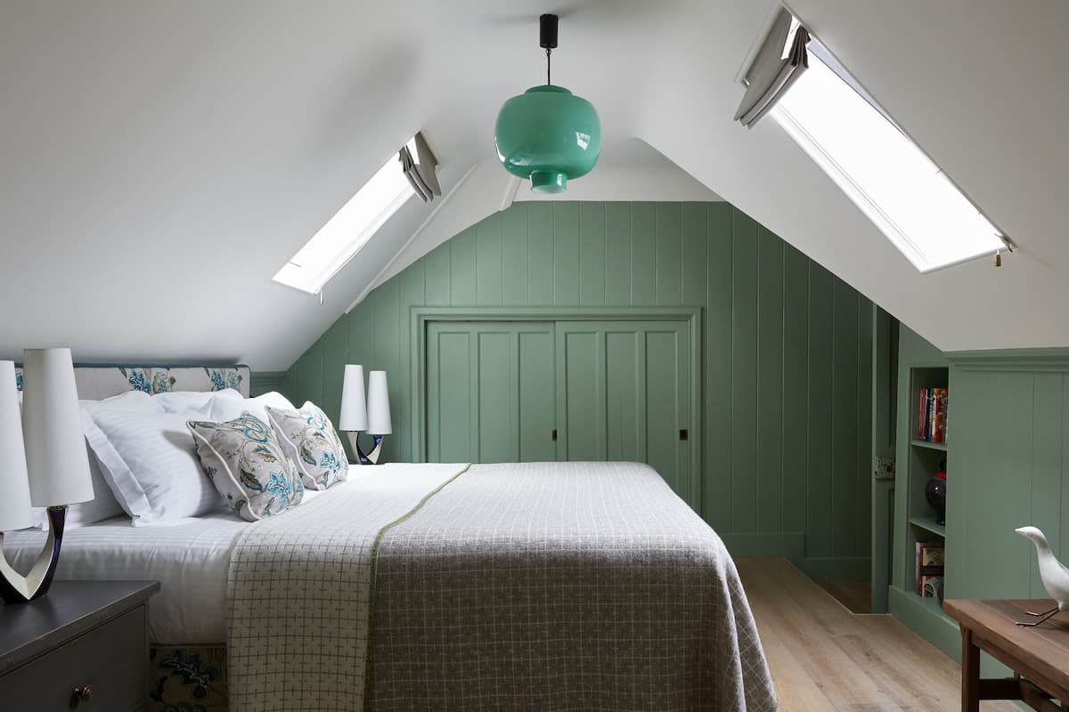 Un dormitorio con cama, lámparas de noche, pared verde, tragaluz y una estantería.