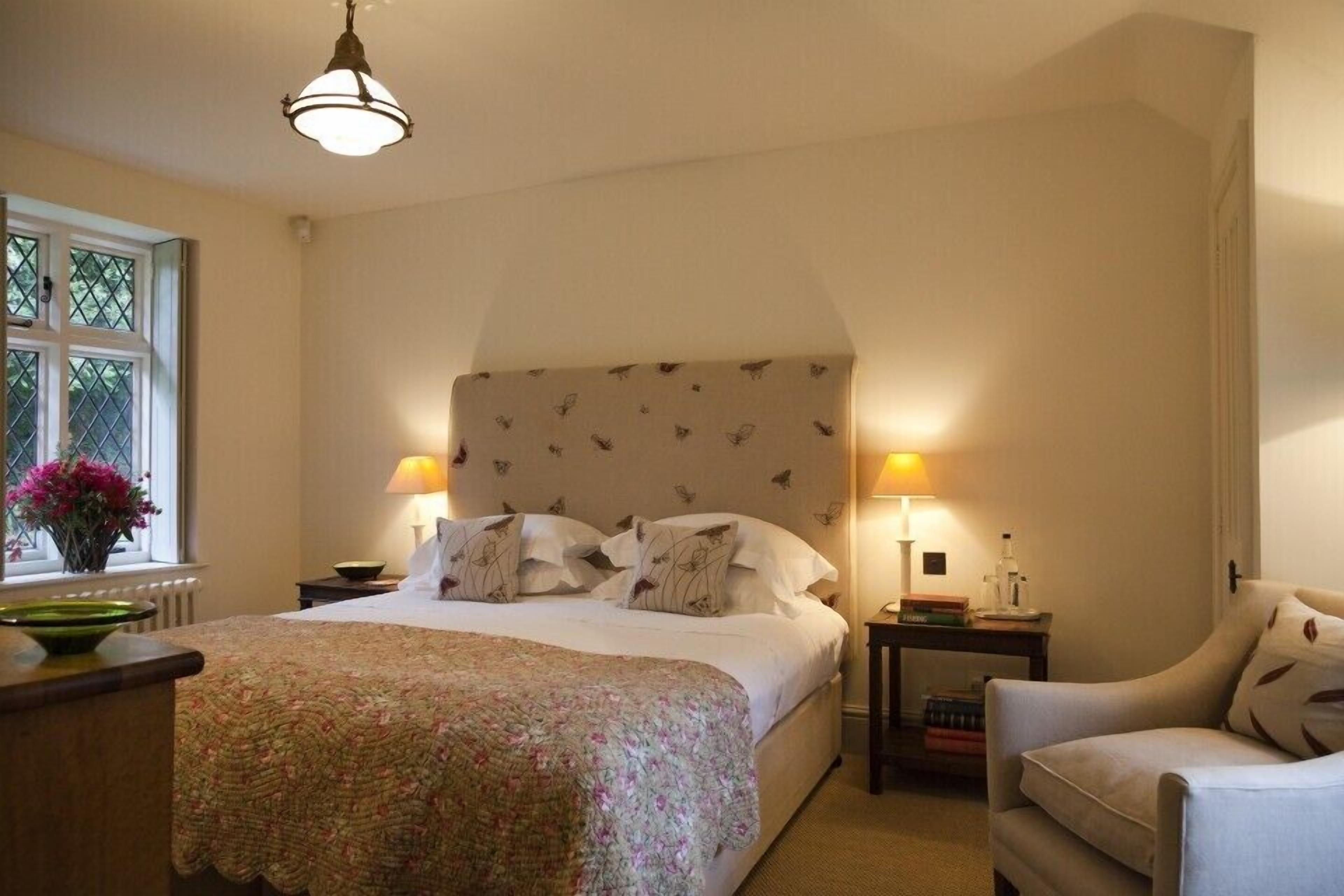Un dormitorio con una cama grande, dos mesitas de noche con lámparas, una silla y una ventana con flores.