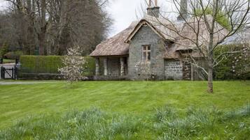 A stone cottage with a thatched roof, surrounded by a well-maintained lawn and trees.