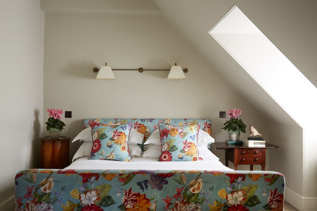Un dormitorio con cama, ropa de cama estampada, una mesita de noche con una planta y una lámpara de pared.