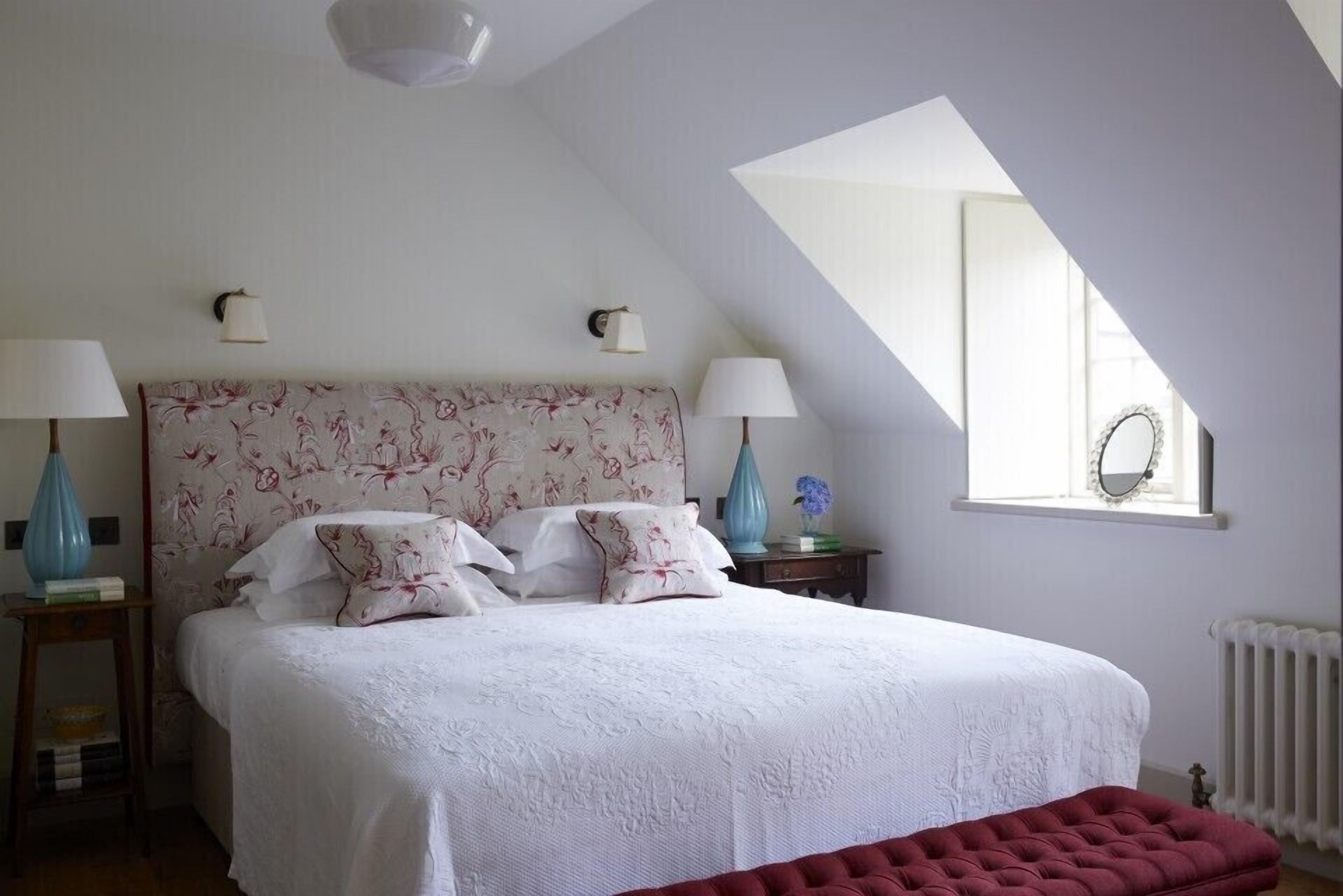 A bedroom with a large bed, two bedside lamps, a red bench, a window with a mirror, and a radiator.