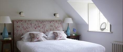 A bedroom with a large bed, two bedside lamps, a red bench, a window with a mirror, and a radiator.