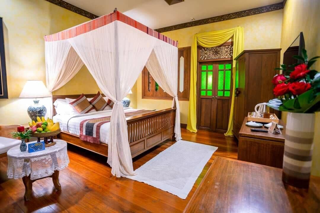 Una cama con dosel y pilares, piso de madera, una mesita redonda con un florero y un aparador de madera.
