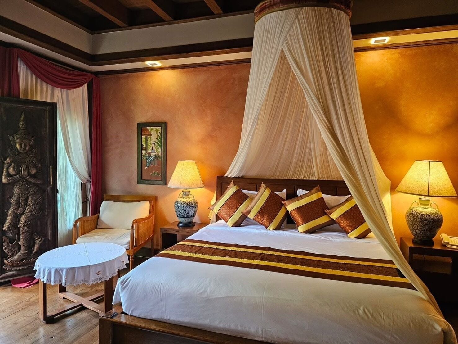 A hotel room with a bed, bedside lamps, a chair, a small table, and a decorative wall piece.