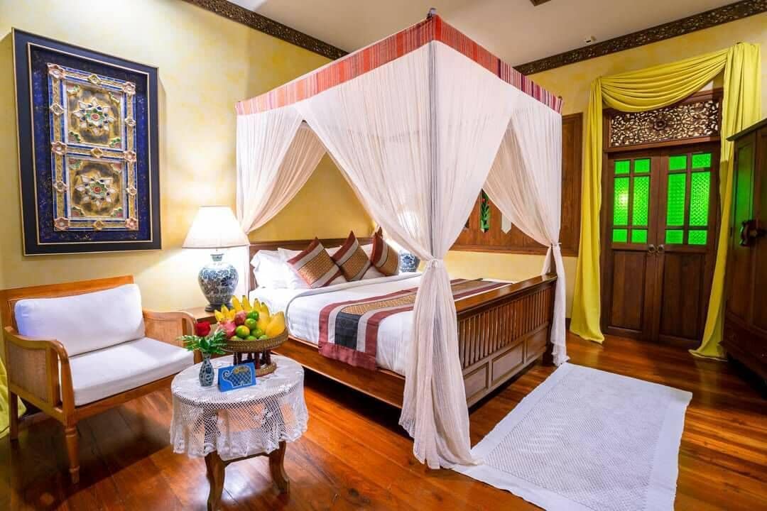 A four-poster bed with a canopy, a wooden bench, a small round table with a vase and fruit, and a green window with decorative shutters.