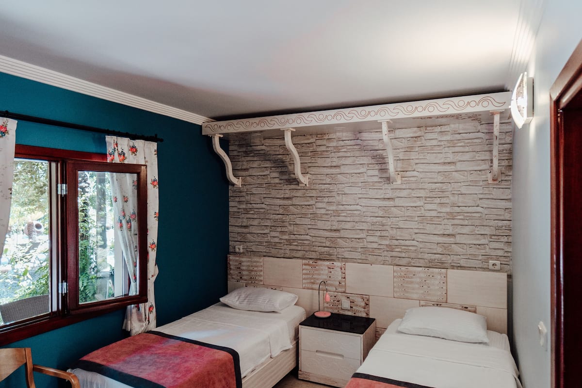 A bedroom with two beds, a window with curtains, a wooden chair, and a brick-patterned wall.