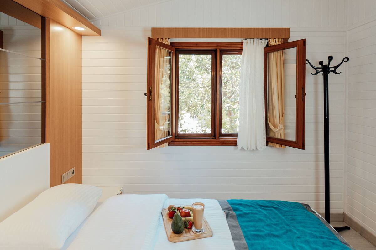 A bedroom with a bed, a window with curtains, a coat rack, and a tray with fruits and a drink.