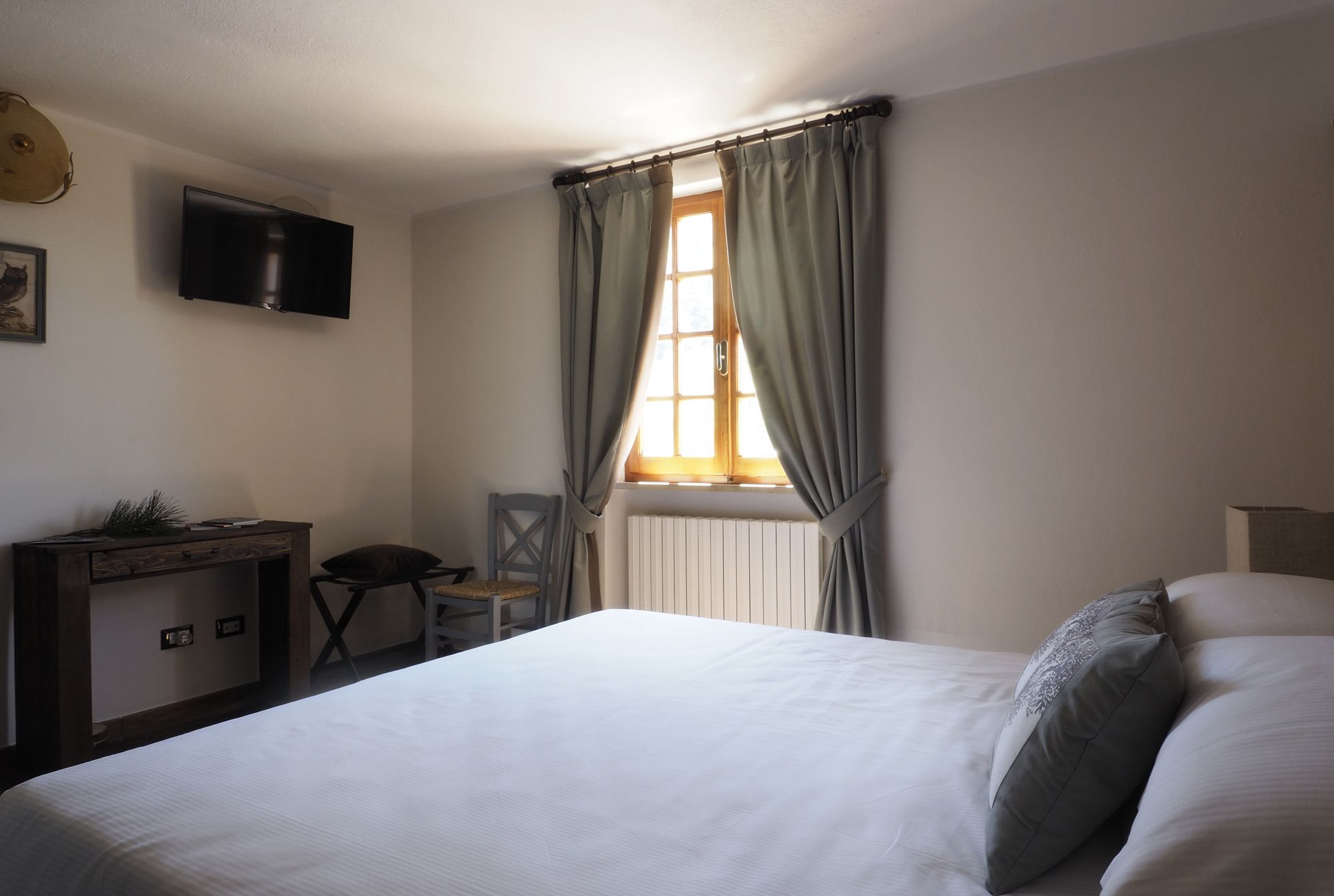 Quarto com cama, escrivaninha, cadeira, televisão e janela com cortinas.