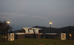 Un espacio al aire libre con mobiliario de mimbre, una botella y vasos, rodeado de faroles y un paisaje montañoso.