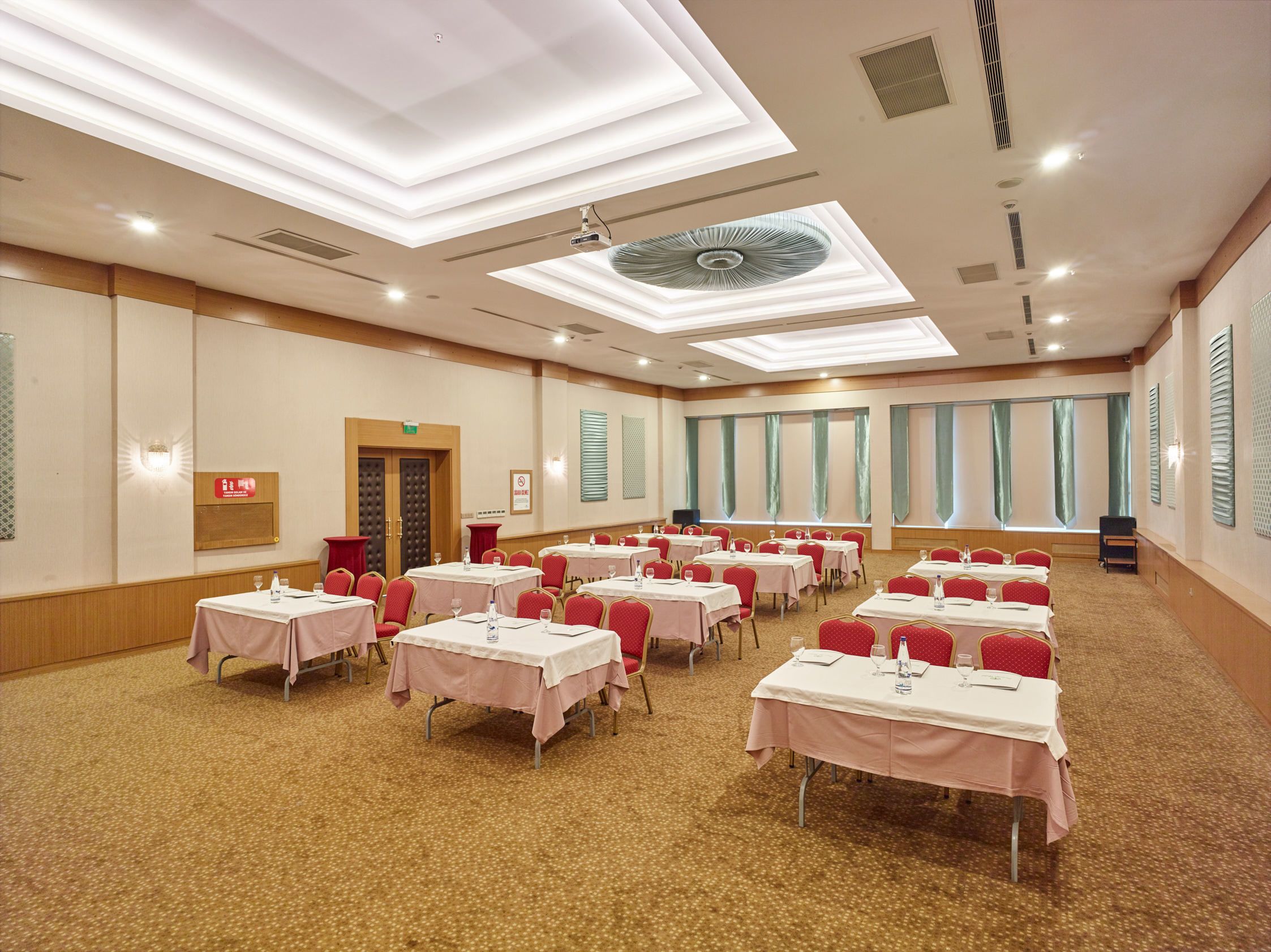 Un salón de conferencias con varias mesas, sillas rojas y un ventilador de techo.