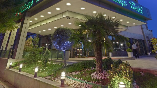 Un hotel con fachada moderna, señalización iluminada y un área de entrada ajardinada.