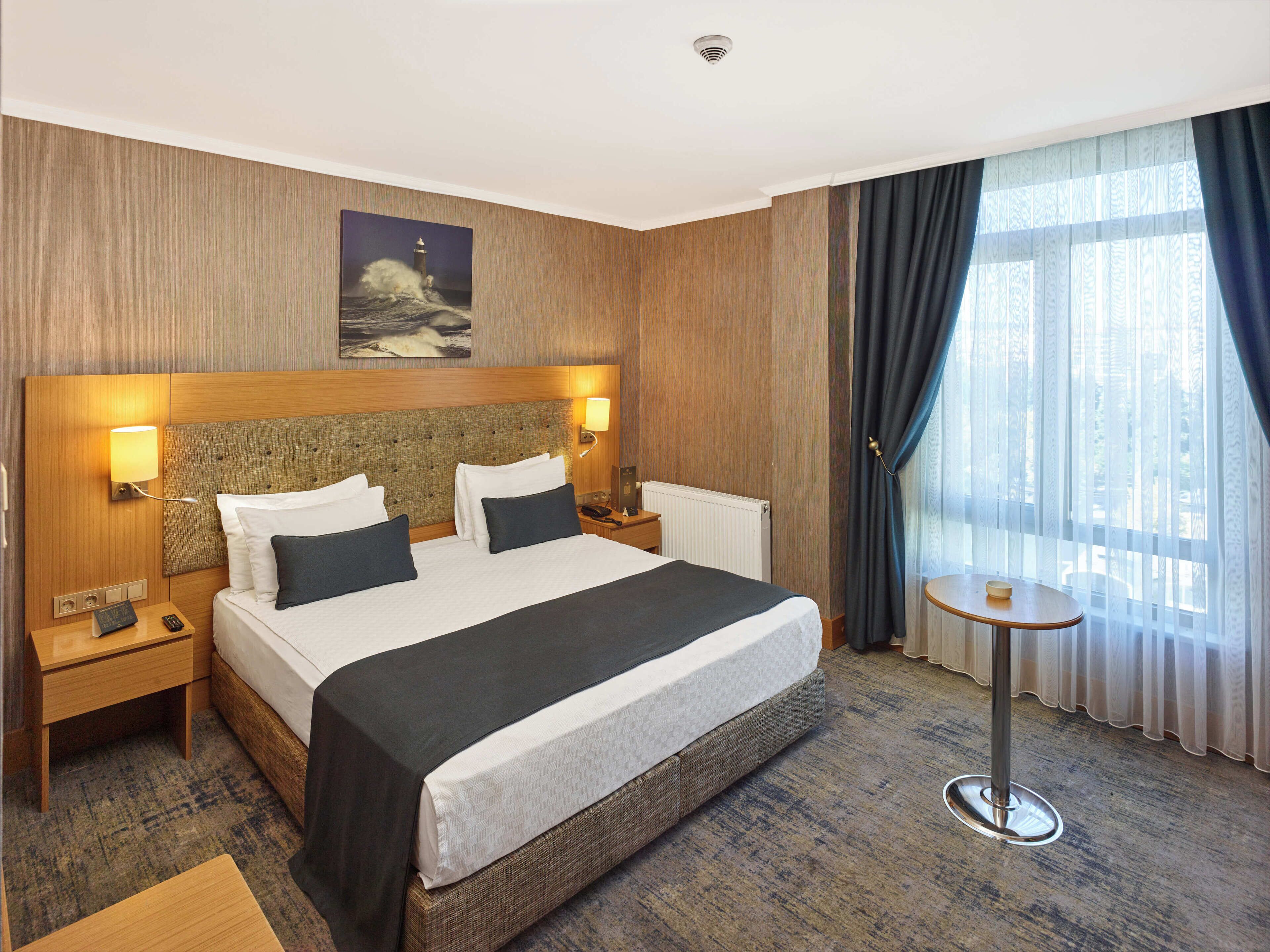 A hotel room with a large bed, two bedside tables, a small table, and a window with curtains.