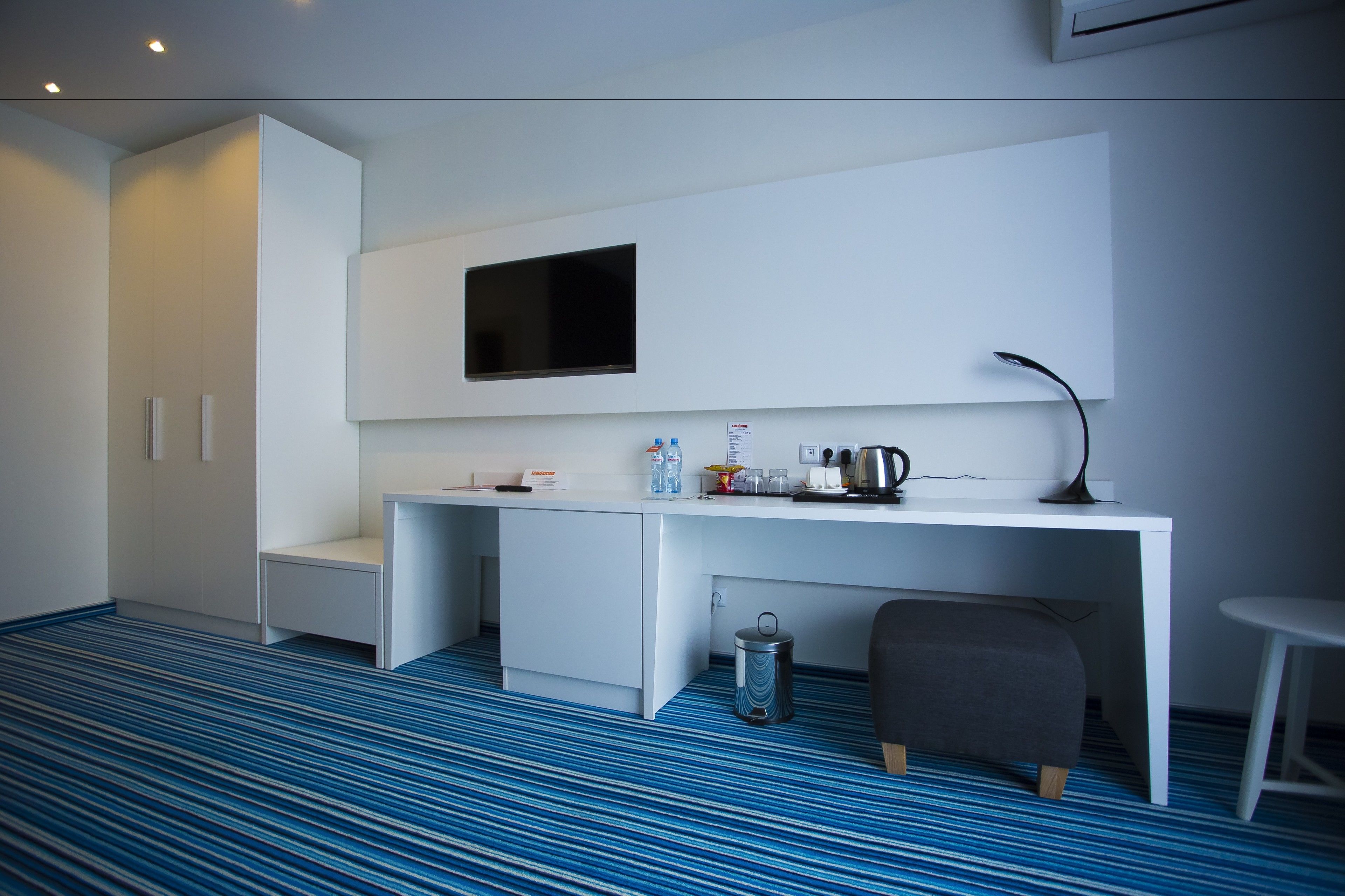A modern hotel room with a white desk, a flat-screen TV, a small trash bin, and a grey ottoman.
