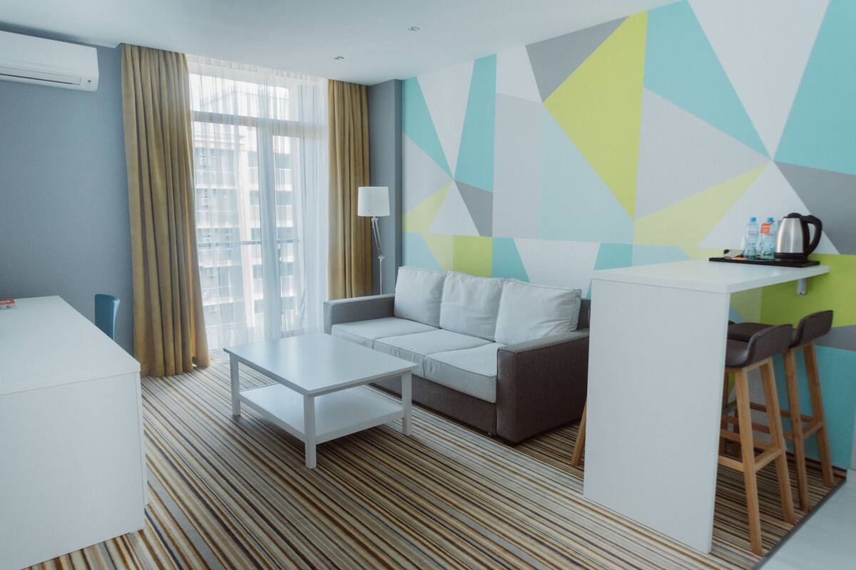 A modern hotel room with a sofa, a small table, a bar area, and a view through the window.
