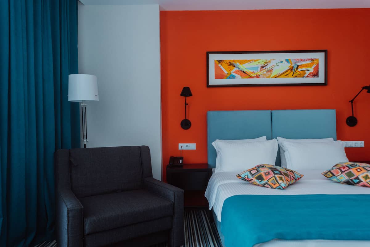 A hotel room with a bed, a chair, a nightstand, a lamp, and a painting on the wall.