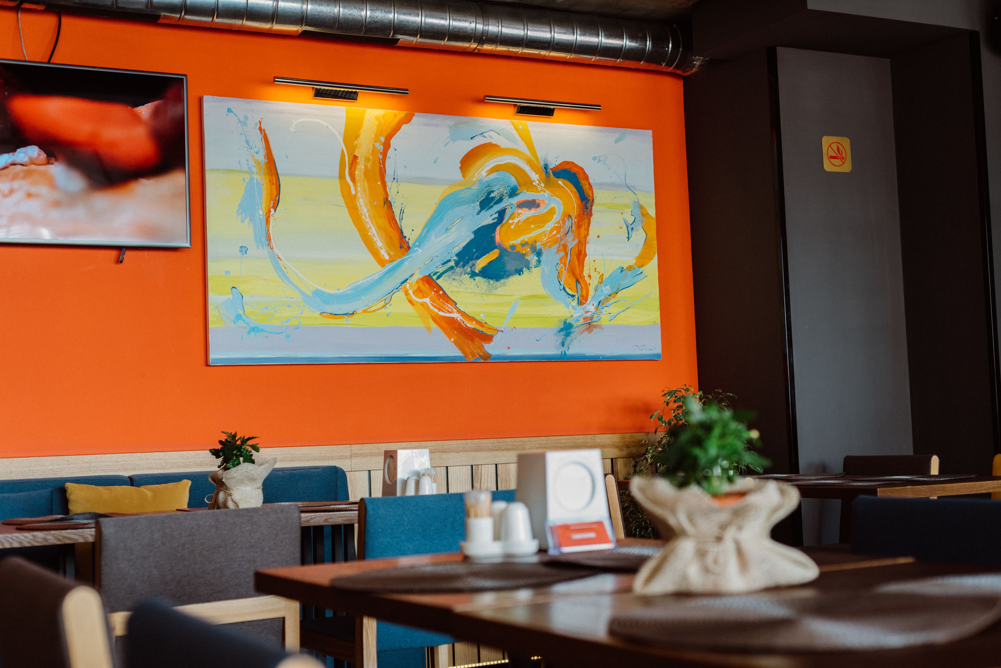 A restaurant interior with a large abstract painting on the wall, wooden tables, and blue and yellow seating.