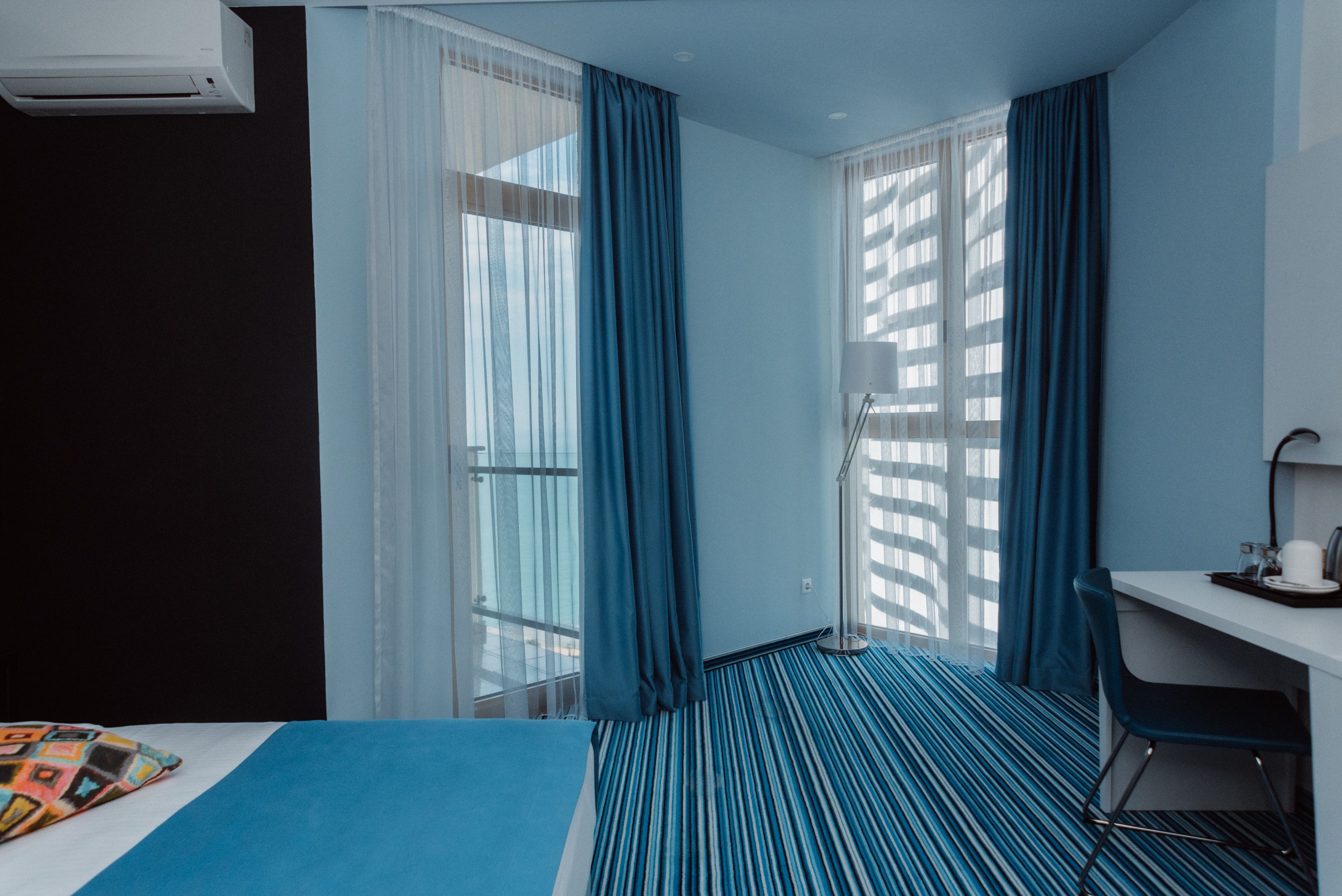 Quarto de hotel com cama, escrivaninha e cadeira. Há uma janela com cortinas e uma luminária na escrivaninha.