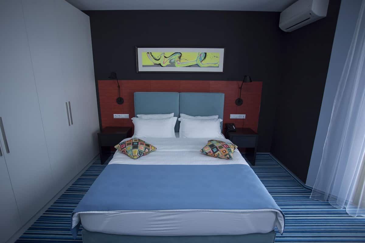 Une chambre d’hôtel avec un lit, une tête de lit, un climatiseur fixé au mur, un tableau encadré et une moquette à rayures.