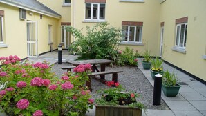 Une cour avec un banc en bois, des plantes en pot et un bâtiment aux murs et aux fenêtres jaunes.