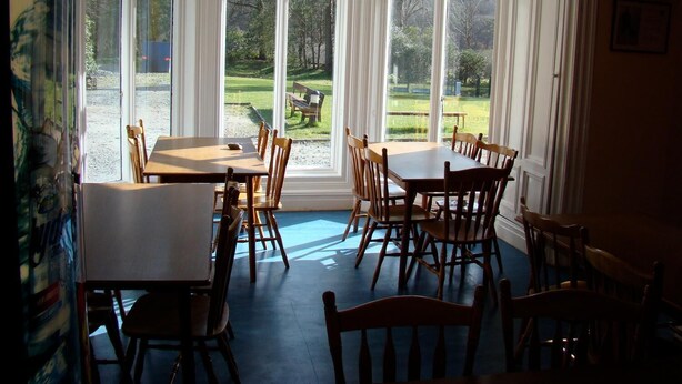 Un espace repas doté de grandes baies vitrées, de tables et de chaises en bois, et offrant une vue sur un espace extérieur verdoyant.