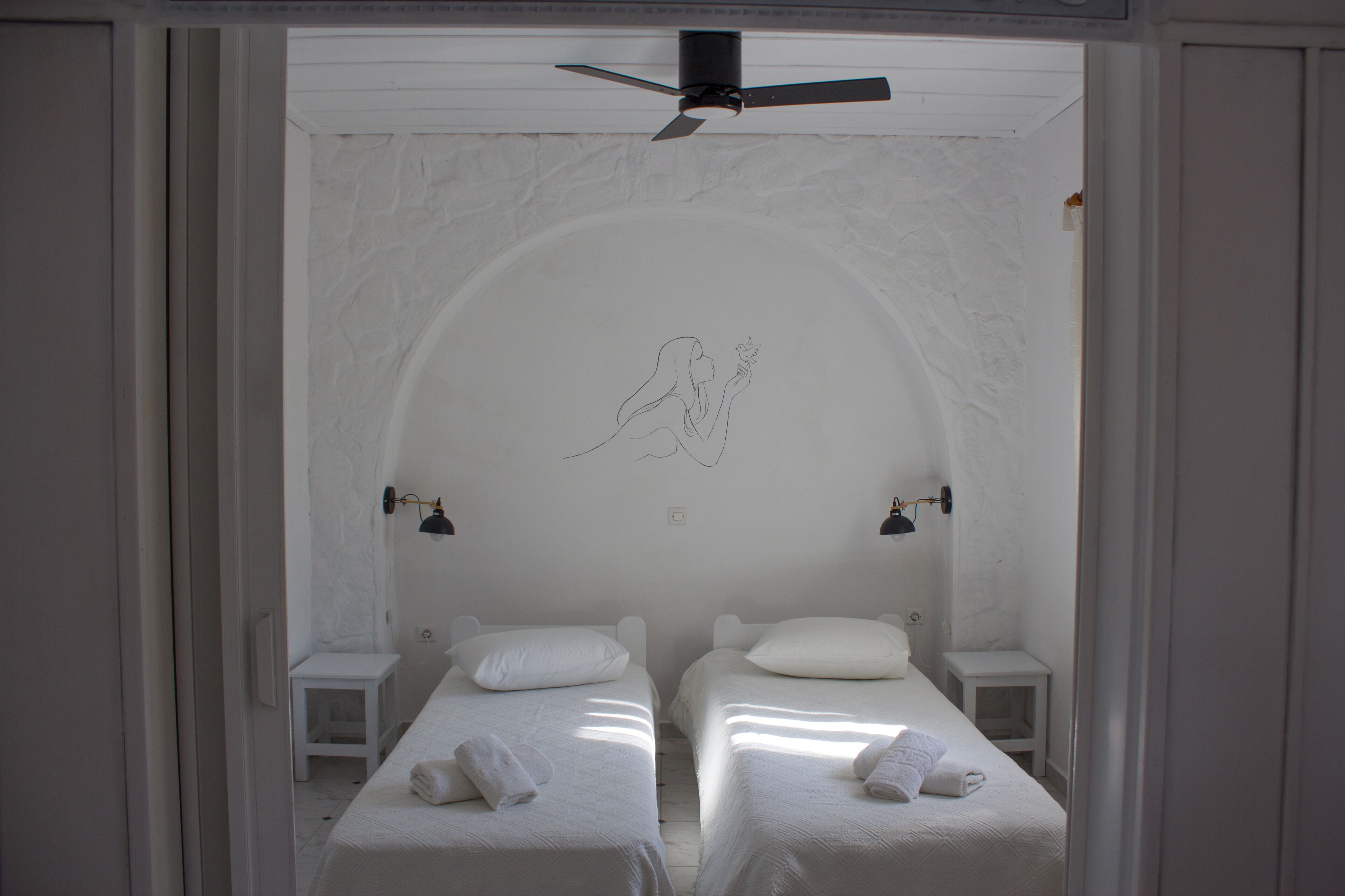 A room with two beds, each with a pillow and a towel. There is a ceiling fan above and a wall with a decorative line drawing.