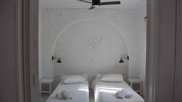 A room with two beds, each with a pillow and a towel. There is a ceiling fan above and a wall with a decorative line drawing.