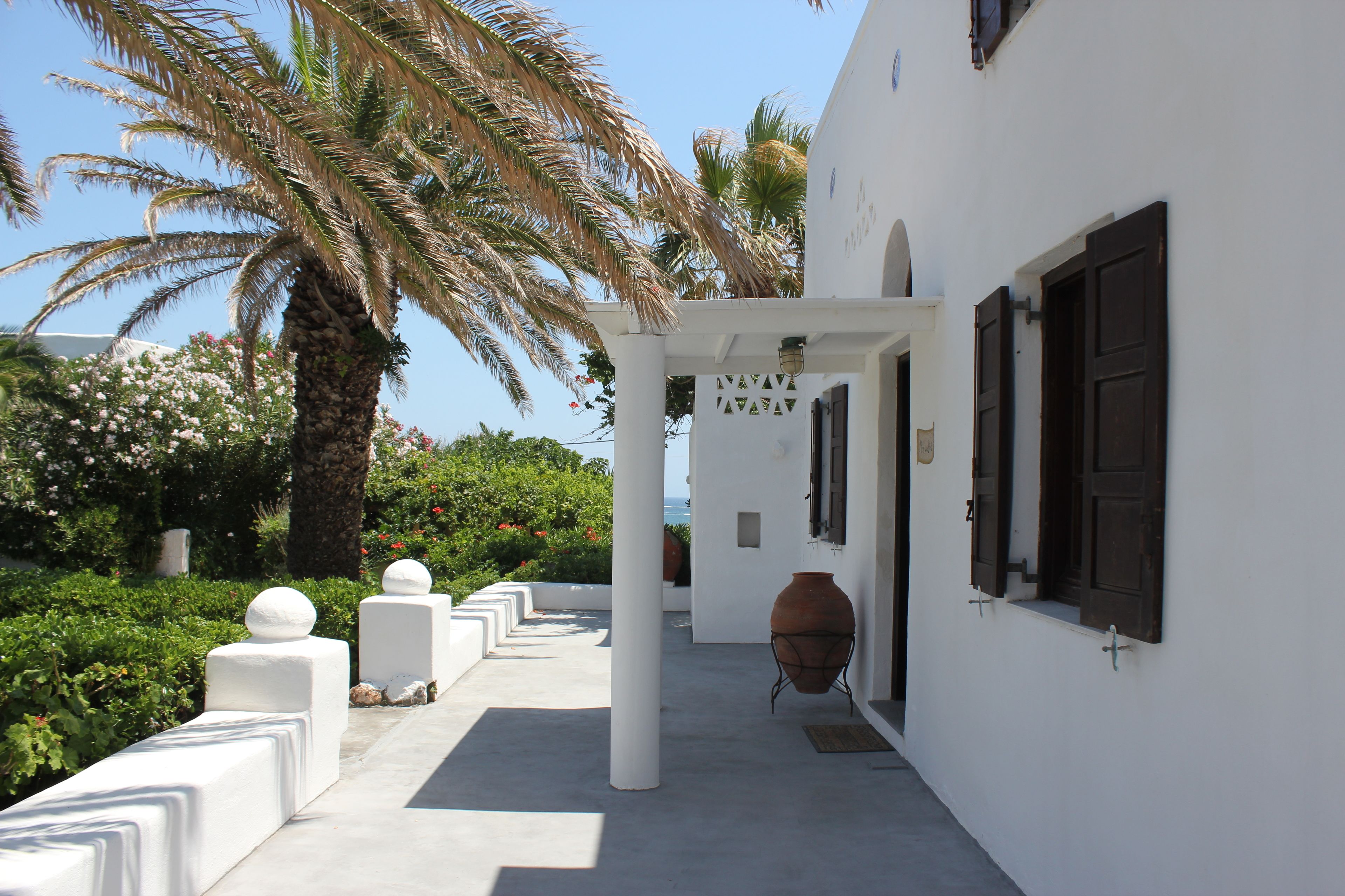A white building with a covered walkway, palm trees, and a large clay pot.