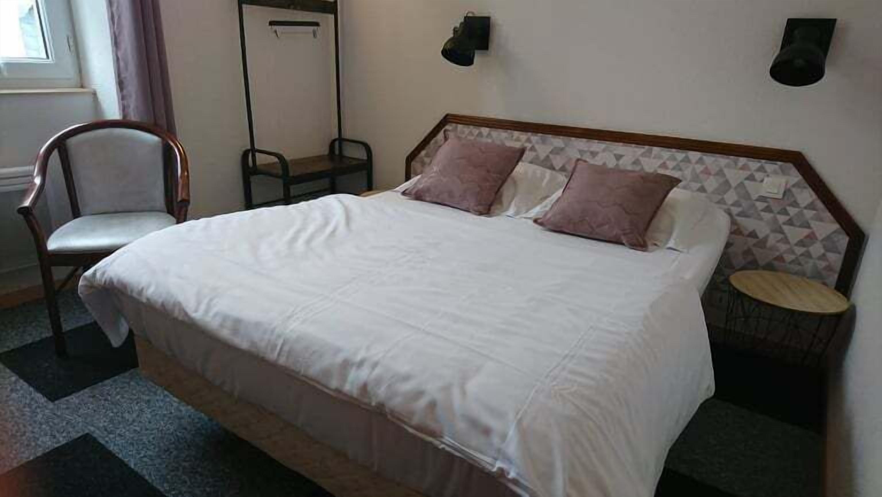 A neatly made bed with white bedding and two pillows, a wooden chair, a small round table, and a wall-mounted light fixture.