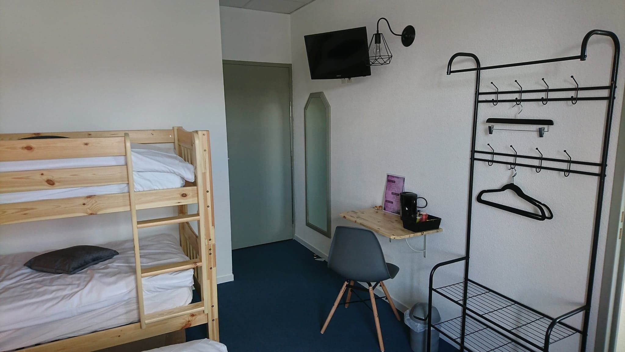 A small, compact room with a bunk bed, a desk with a chair, a TV mounted on the wall, and a coat rack with hangers.