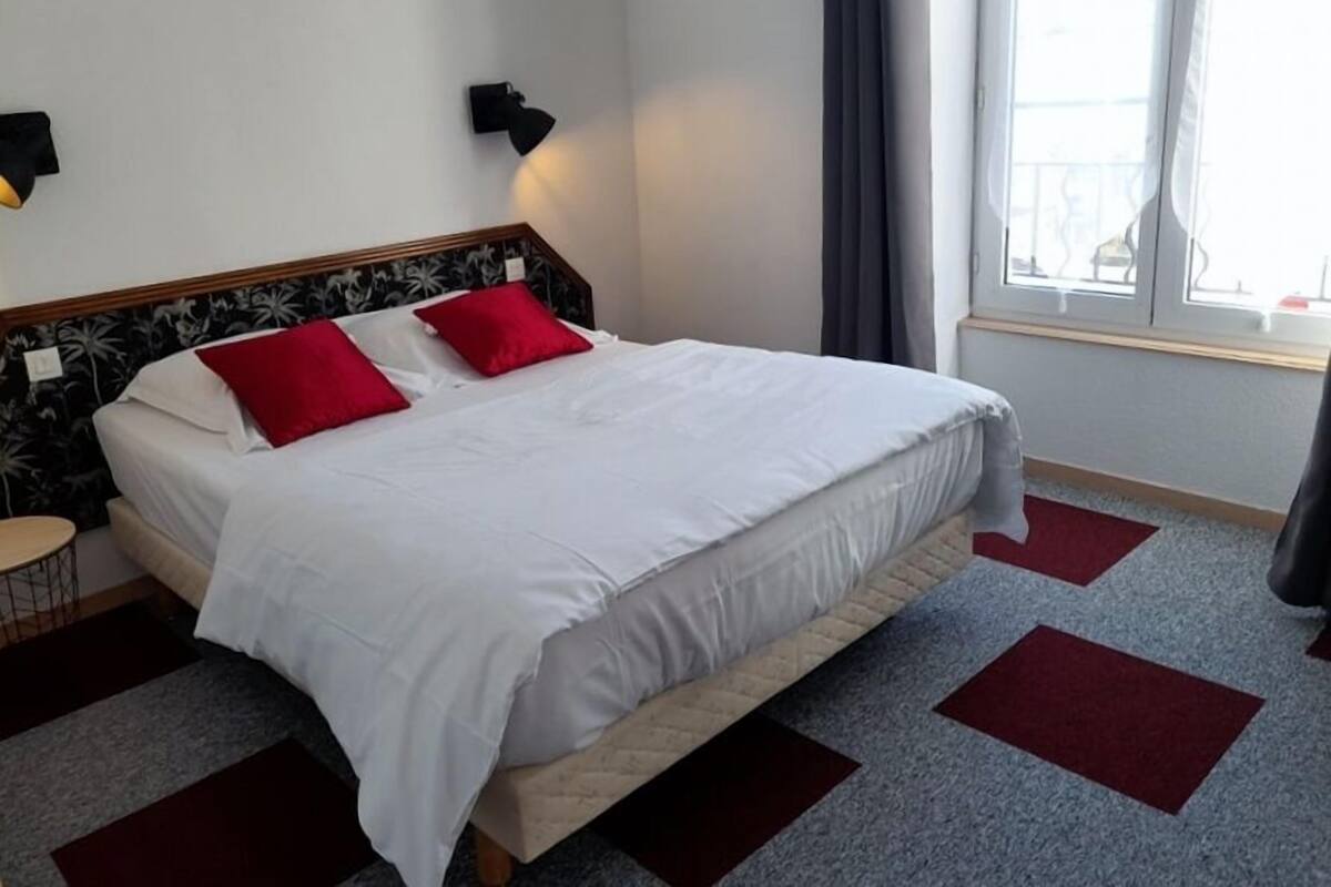 A neatly made bed with white bedding and red pillows, a wooden headboard with a floral pattern, a small bedside table, and a window with curtains.