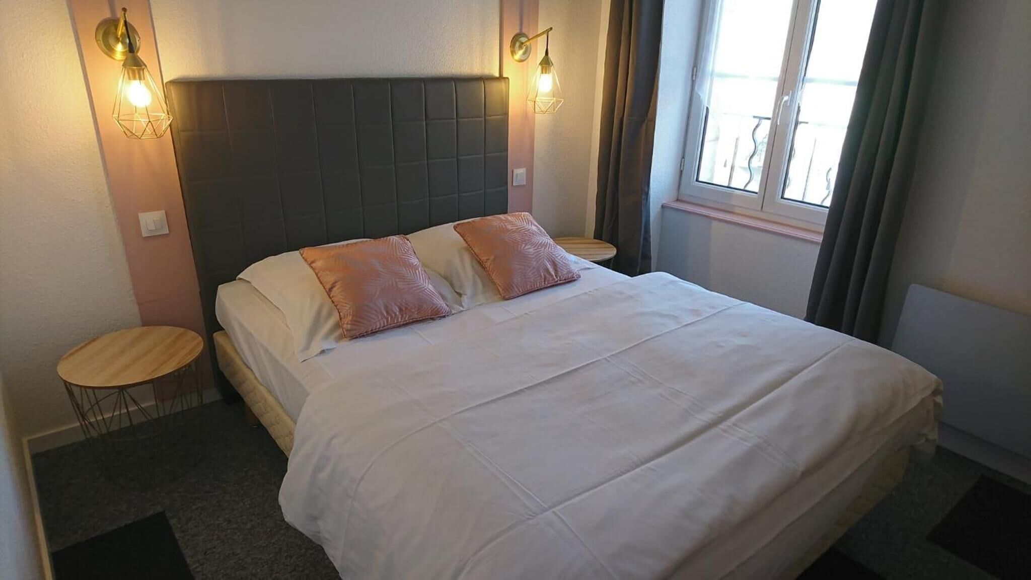 A neatly made bed with two pillows, a headboard, two bedside tables, and a window with curtains.