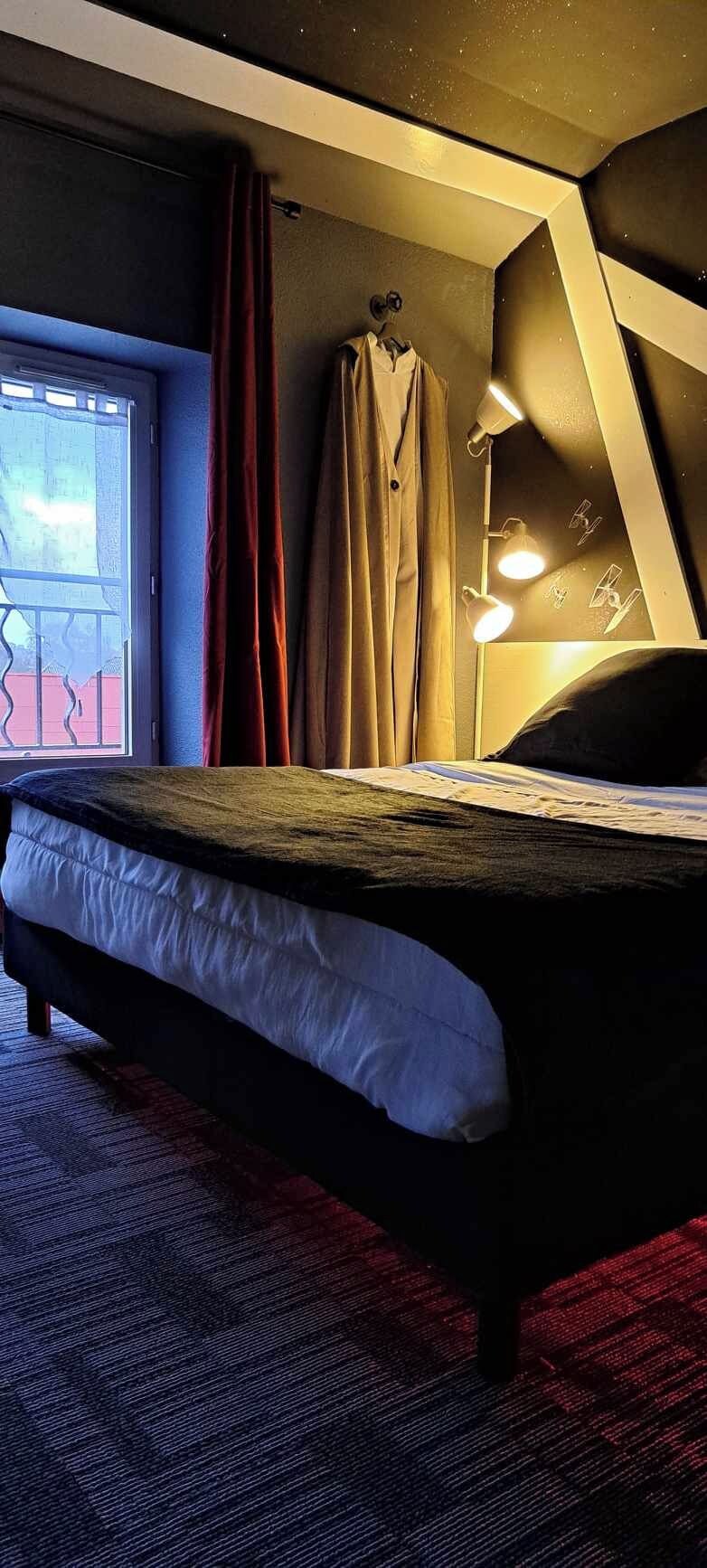Ein Schlafzimmer mit Bett, Vorhängen am Fenster, einem aufhängbaren Mantel und einer Nachttischlampe.