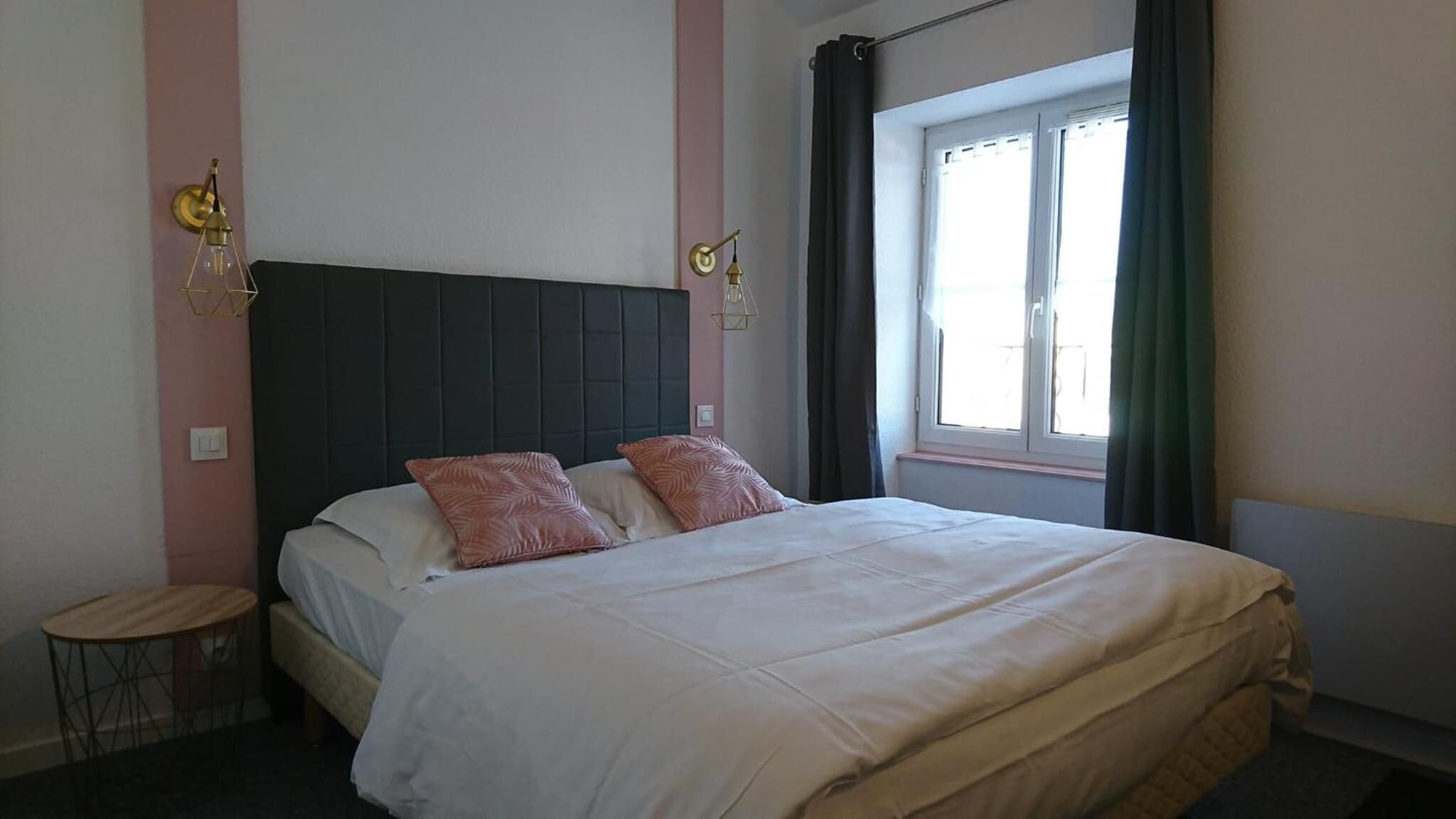 A bedroom with a bed, two wall sconces, a small round table, and a window with curtains.