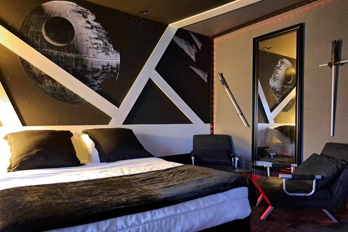 A modern hotel room with a large bed, a desk, and a chair. There is a wall with a space-themed mural.